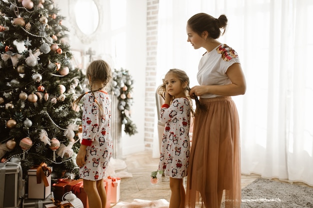 Madre cariñosa trenza la trenza de su pequeña hija mientras la segunda hija decora un árbol de Año Nuevo en la habitación luminosa y acogedora.