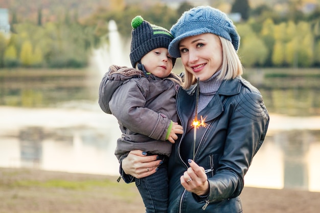 Madre con una chaqueta abrigada y gorro de punto abrazando a su bebé caminando en el parque de otoño. Elegante mujer rubia con un niño con el telón de fondo del lago