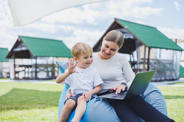Una madre con una computadora portátil, su hijo sentado junto a ella al aire libre en un césped verde Descansa y trabaja con un niño