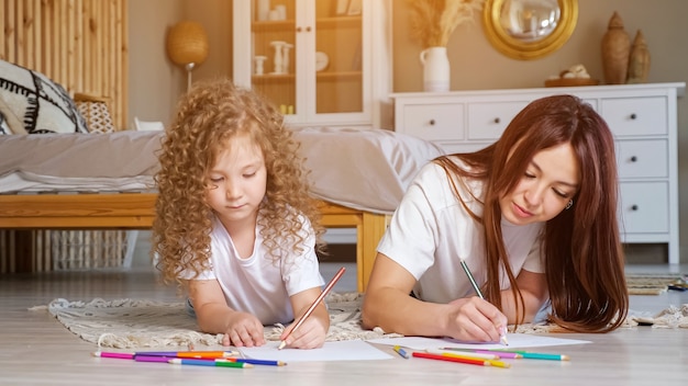 Madre concentrada e hija en edad preescolar con cabello largo suelto dibujar en papeles con lápices de colores tirado en el piso en casa, luz del sol