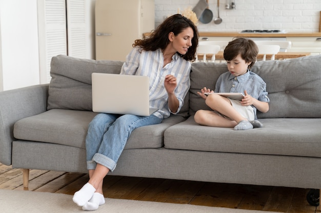 Madre curiosa mira la tableta como hijo en edad preescolar, juega a juegos, trabajador remoto, mamá, trabaja en una computadora portátil con un niño