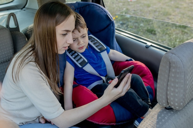 La madre le da el teléfono al niño pequeño sentado en un asiento de seguridad. Seguridad del transporte de niños.