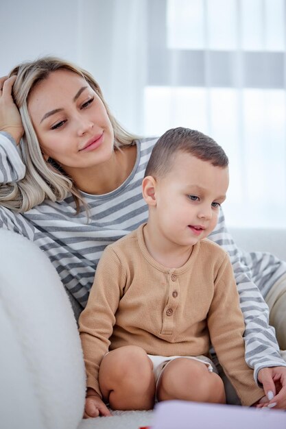 Madre despreocupada descansar con su hijo en edad preescolar en la sala de estar viendo dibujos animados, en casa. Actividad alegre y divertida de la madre y el niño de la familia para el fin de semana en interiores o relajarse en cuarentena