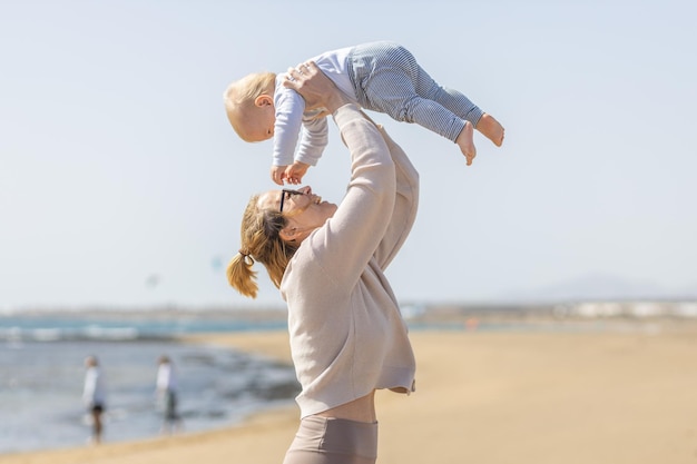 Madre disfrutando de las vacaciones de verano jugando y levantando a su hijo pequeño en el aire en la playa de arena en la isla de Lanzarote España Concepto de viajes y vacaciones familiares