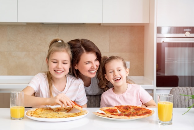 Madre y dos hijas comiendo pizza casera en una mesa en la cocina, concepto de familia feliz