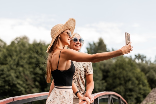 Madre de dos mujeres felices con una hija sonriente en un traje de verano de moda con gafas de sol vintage descansan juntos en la naturaleza y toman una foto de grupo en el teléfono