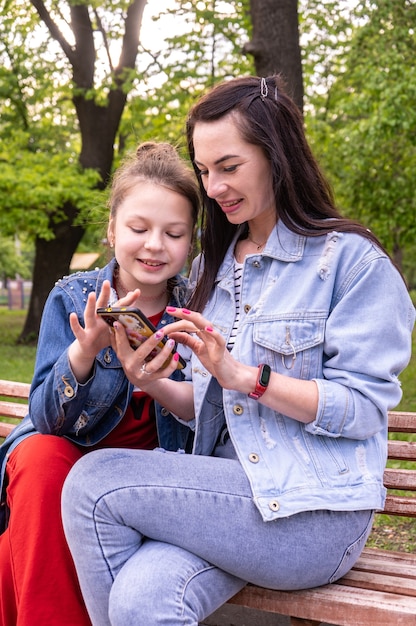 Madre e hija adolescente caminando en un parque, sentada en un banco y mirando en el teléfono inteligente, feliz joven caucásica con cabello largo y adolescente navegando por internet al aire libre, estilo de vida familiar