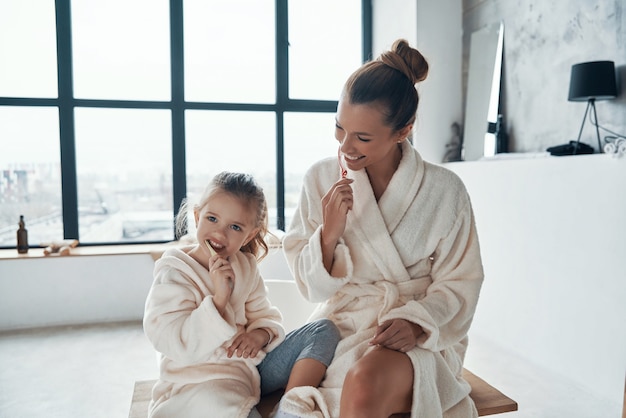 Madre e hija en albornoces sonriendo y limpiando los dientes mientras hacen la rutina matutina