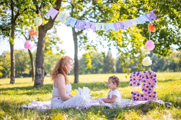 Madre e hija alegres que se divierten en el cumpleaños del niño en una manta con adornos de papel en el parque