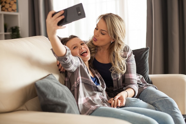 Madre e hija alegres sentadas en el sofá de la sala de estar tomando un selfie juntos.