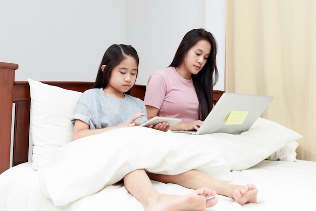 Madre e hija asiáticas mirando portátil y tableta en la cama en el dormitorio