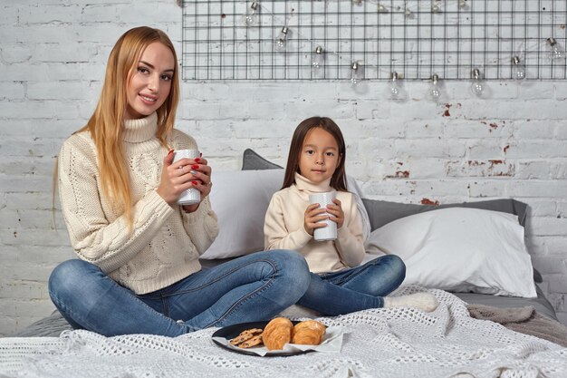 Madre e hija beben té o cacao de tazas grandes sentadas en la cama