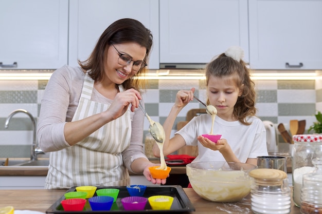 Madre e hija cocinando muffins juntos en la cocina de casa. Mujer y niño vertiendo masa cruda en moldes de silicona. Día de la madre, familia, horneado casero de alimentos saludables.