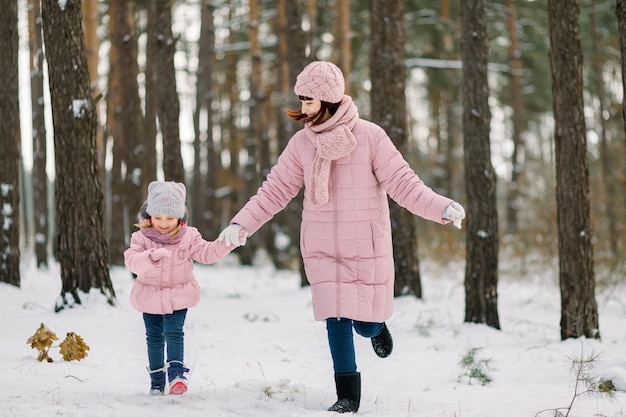 Madre e hija se divierten y se toman de la mano mientras corren y ríen en el bosque nevado de invierno entre los hermosos pinos. Familia feliz disfrutando de tiempo juntos.