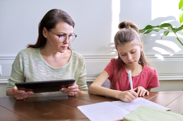 Madre e hija estudian juntos en casa, sentados a la mesa. Mujer con tableta digital, utilizando Internet, niña escribiendo en el cuaderno. Aprendizaje a distancia, padres ayudando a su hijo a un estudiante de escuela primaria