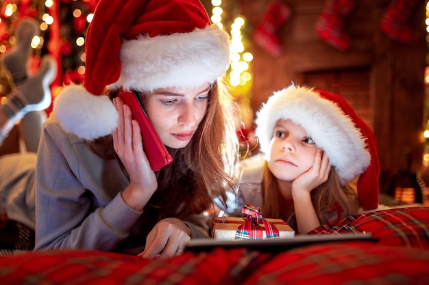 Madre e hija de la familia con sombreros y pijamas eligiendo regalos en tableta digital y mamá llama a Papá Noel por teléfono móvil