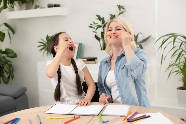 Madre e hija hermosas y felices hacen una tarjeta para el día de la madre sentadas juntas en la mesa con papel y lápices.