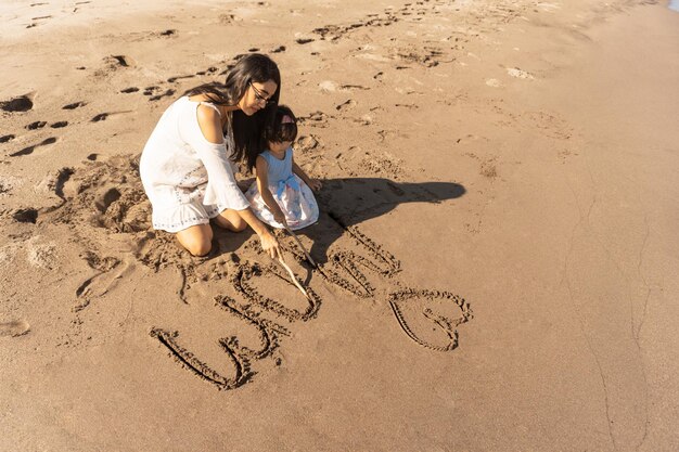 Madre e hija juntas escribiendo "Te amo mamá" en la arena de una playa