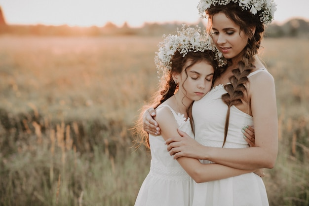 Madre e hija juntas en vestidos blancos con trenzas y coronas florales en estilo boho en el campo de verano al atardecer