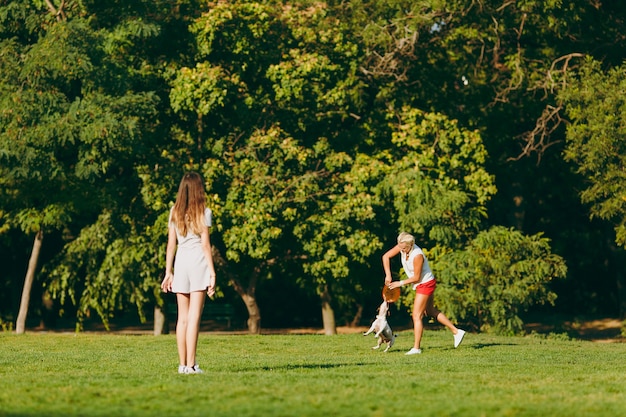 Madre e hija lanzando un disco volador naranja a un pequeño perro gracioso, que lo atrapa en la hierba verde. Pequeña mascota Jack Russel Terrier jugando al aire libre en el parque. Perro y mujer. Familia descansando al aire libre