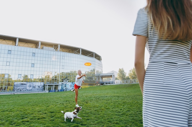 Madre e hija lanzando un disco volador naranja a un pequeño perro gracioso, que lo atrapa en la hierba verde. Pequeña mascota Jack Russel Terrier jugando al aire libre en el parque. Perro y mujer. Familia descansando al aire libre