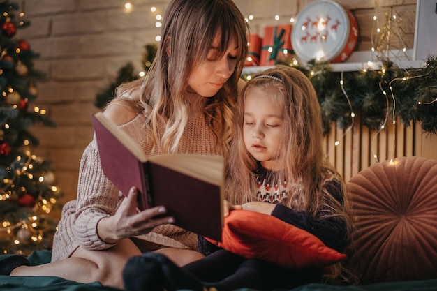 Madre e hija leyendo cuentos de hadas durante la víspera de Navidad, maravillosa atmósfera de amor y confianza, creencia en milagros, felices fiestas