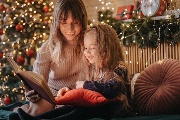 Madre e hija leyendo cuentos de hadas durante la víspera de Navidad, maravillosa atmósfera de amor y confianza, creencia en milagros, felices fiestas