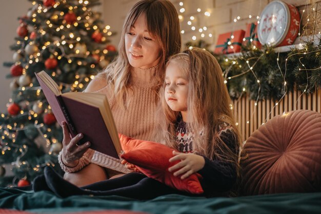 Madre e hija leyendo cuentos de hadas durante la víspera de Navidad, maravillosa atmósfera de amor y confianza, creencia en milagros, felices fiestas