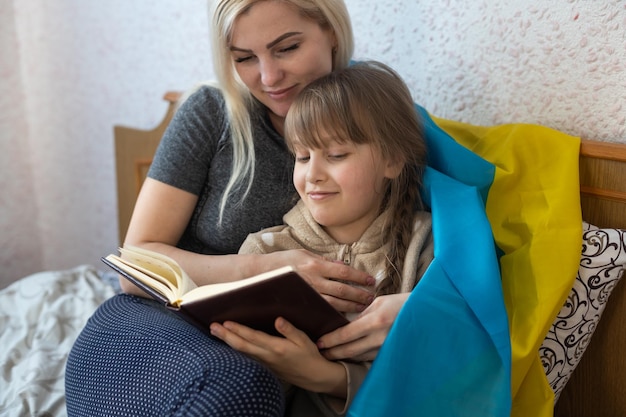 Madre e hija leyendo un libro con la bandera de ucrania en la cama