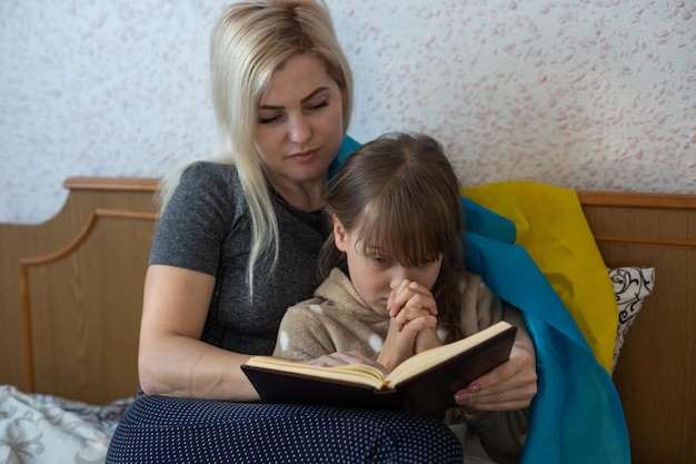 Madre e hija leyendo un libro con la bandera de ucrania en la cama