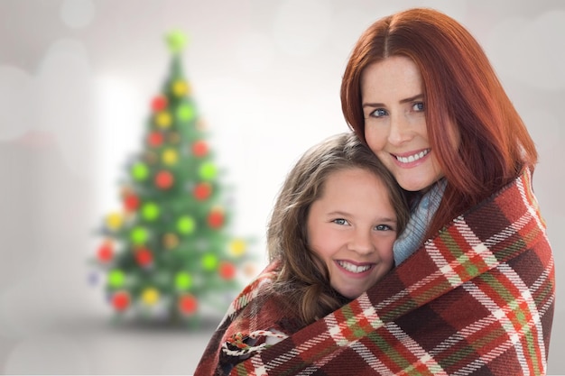 Madre e hija bajo una manta contra un árbol de navidad borroso en la habitación