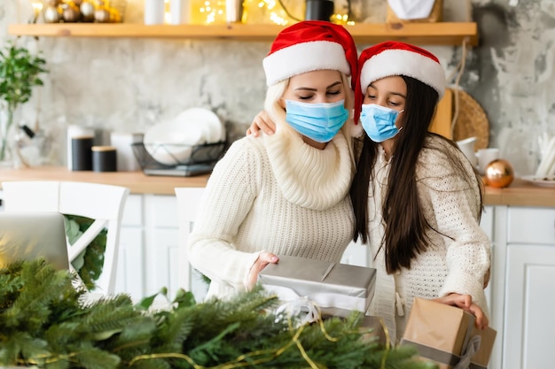 madre e hija con máscaras médicas bajo el árbol de navidad durante la celebración en casa.