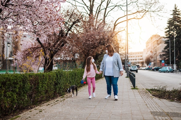 Madre e hija con máscaras protectoras en sus rostros disfrutando de un paseo en el parque con su perro.