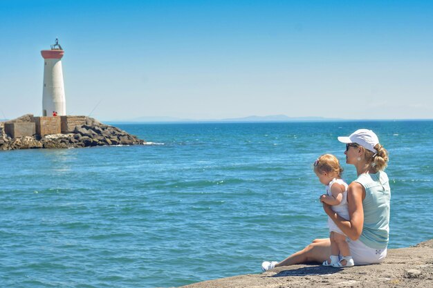 Madre e hija en el muelle de la costa mediterránea con faro