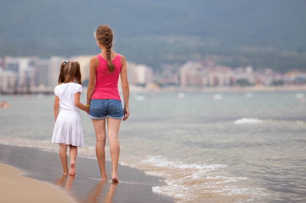 Madre e hija niña caminando juntos en la playa de arena en agua de mar en verano con los pies descalzos en las cálidas olas del océano.