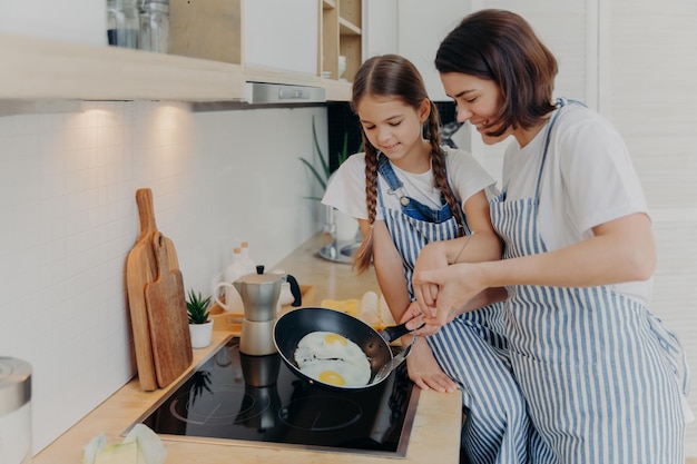 Madre e hija ocupadas usan delantales a rayas posan en la cocina cerca de la cocina freír huevos en la sartén preparar un desayuno rápido disfrutar de un ambiente doméstico Mamá enseña a un niño pequeño a cocinar Concepto de familia feliz