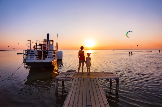 Madre e hija de pie en el muelle en el fondo del mar, barco y puesta de sol. Paisaje ucraniano