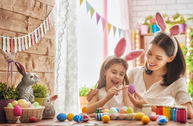 Madre e hija pintando huevos. Familia feliz preparándose para la Pascua. Niña linda con orejas de conejo.