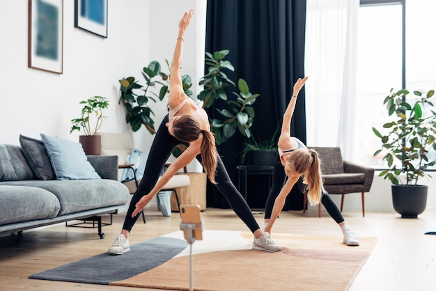 Madre e hija practicando en casa haciendo ejercicio de molino de viento girando el torso doblado y tocando la pierna con las manos