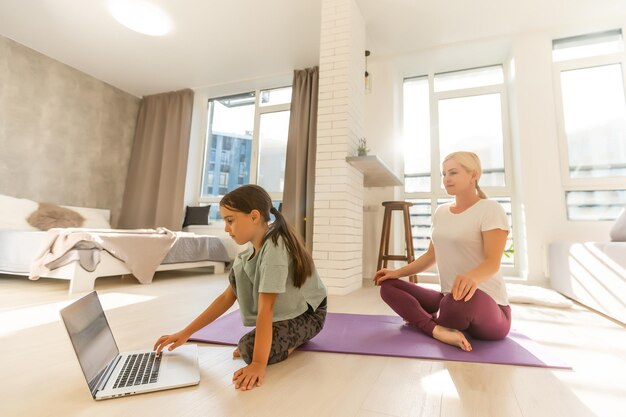 Madre e hija practicando clases de yoga en línea en casa durante la cuarentena durante la pandemia del coronavirus. Familia haciendo deportes juntos en línea desde casa durante el aislamiento juntos en casa