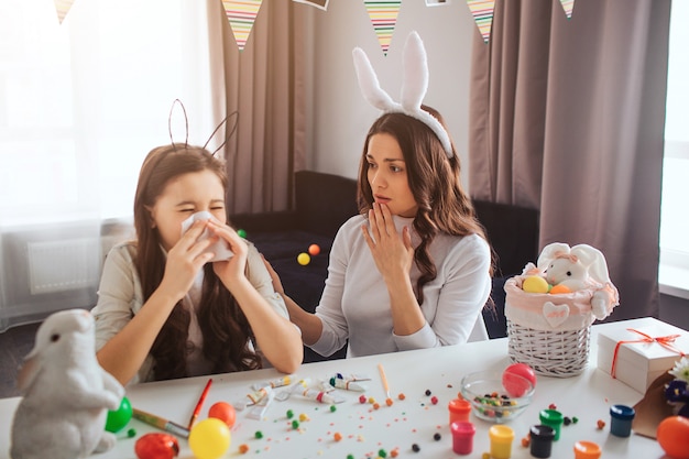 Madre e hija se preparan para la Pascua juntas. Niña enferma estornudando. Mujer joven preocupada. Decoración de Pascua con pintura y dulces en la mesa.