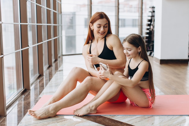 Madre e hija se relajan después del entrenamiento sentado en la estera, usando el teléfono en el gimnasio para ver un video, ¿cómo hacer ejercicio?