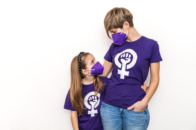 Foto madre e hija rubia abrazándose con camiseta morada con el símbolo del día internacional de la mujer trabajadora feminista en una pared blanca, 8 de marzo, y con una máscara por el coronavirus.