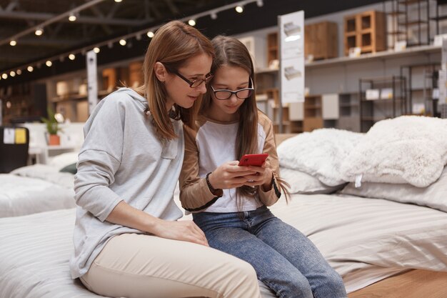 Madre e hija sentada ona cama en la tienda de muebles, usando teléfonos inteligentes juntos