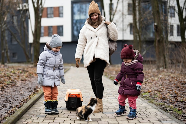 Madre e hijas caminando con gatito en carro de jaula de plástico de viaje al aire libre en el parque