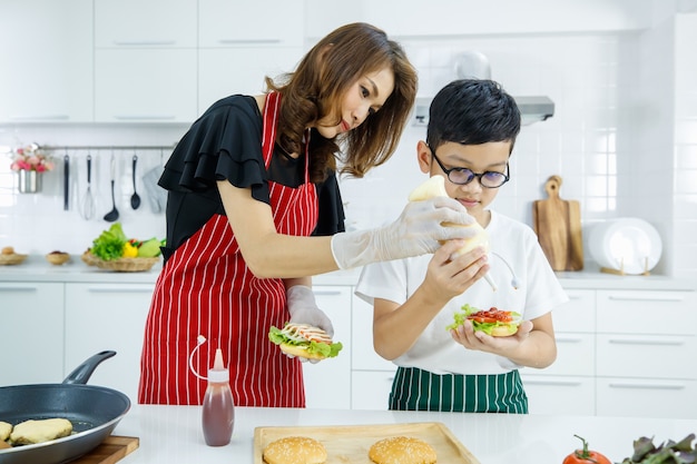 Madre e hijo asiáticos que agregan mayonesa a una hamburguesa fresca mientras preparan el almuerzo en un almuerzo moderno en la cocina de casa. Aprender haciendo el concepto de niño moderno.