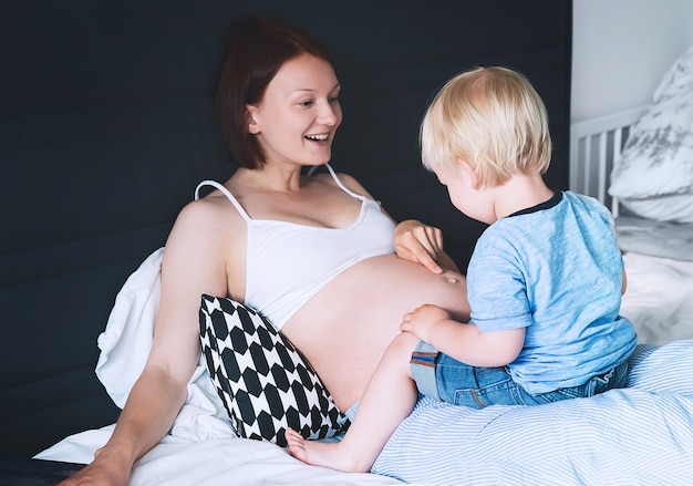 Madre e hijo embarazadas juntos en la cama en casa Niño niño mirando a la barriga de su madre