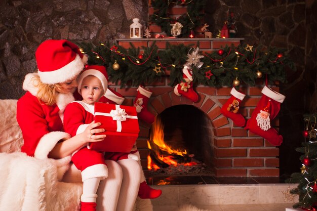 Madre e hijo están sentados junto a la chimenea y el árbol de Navidad. Familia mira en una caja de regalo.