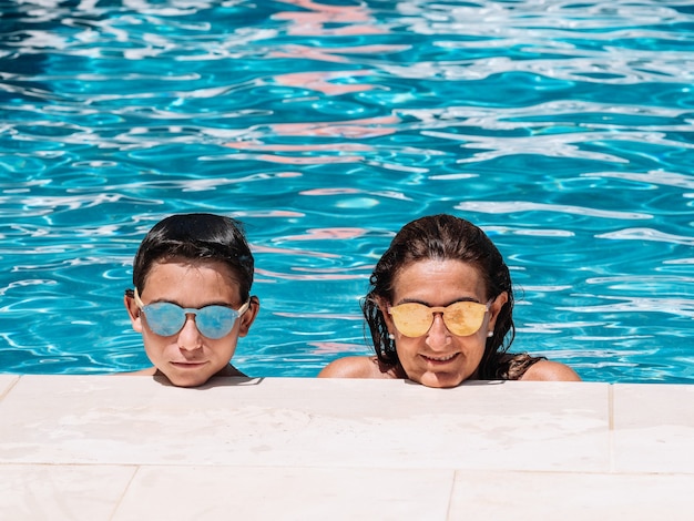 Madre e hijo con gafas de sol están apoyados en el borde de la piscina en un día soleado de verano