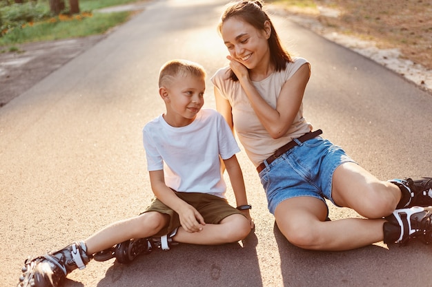 Madre e hijo en patines sentados en una carretera, mamá hablando con su niño, disfrutando de la hermosa naturaleza y el aire fresco, fines de semana activos juntos.